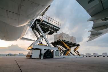 Ein Flugzeug wird auf einem Flugfeld mit Fracht beladen. Der Fotografierende steht dabei unter dem Flugzeug, links oben ist der Bauch und rechts oben der Flügel des Flugzeuges zu erkennen.