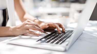 Eine Frau arbeitet an einem Laptop, es sind nur ihre Arme und das weiße Laptop zu sehen. Der Fokus liegt auf Ihren Händen und der Tastatur, die helle Umgebung des Außenbereichs ist leicht unscharf.