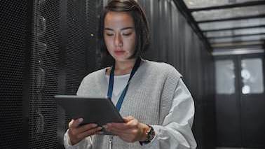 Eine junge dunkelhaarige Frau schaut auf ein Tablet in ihrer Hand, sie steht neben einer Reihe schwarzer Server in einem leicht durch ein Oberlicht beleuchteten Raum.